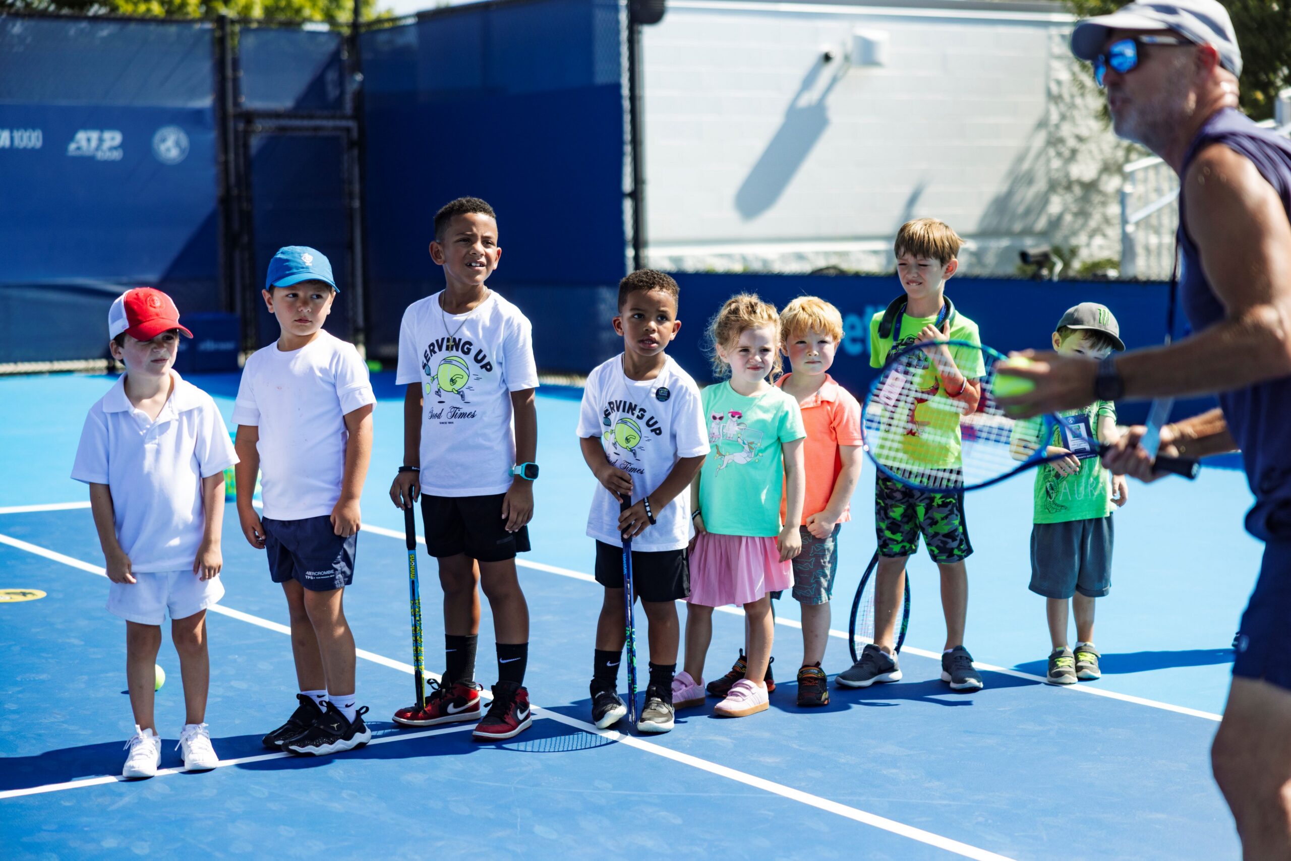 Line of children listening to an adult speak at a tennis clinic.