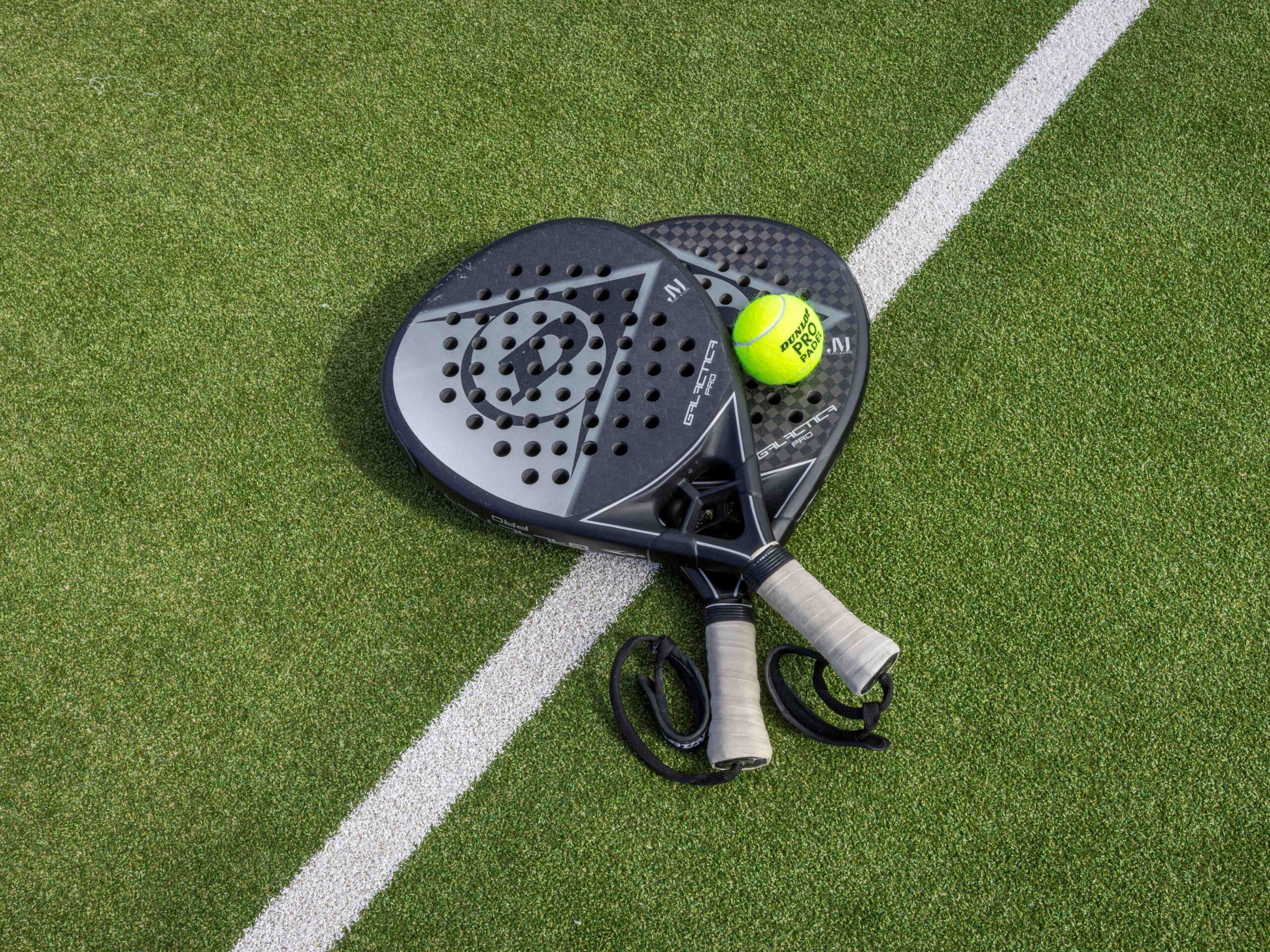 Two Padel racquets sitting on turf with a tennis ball on top